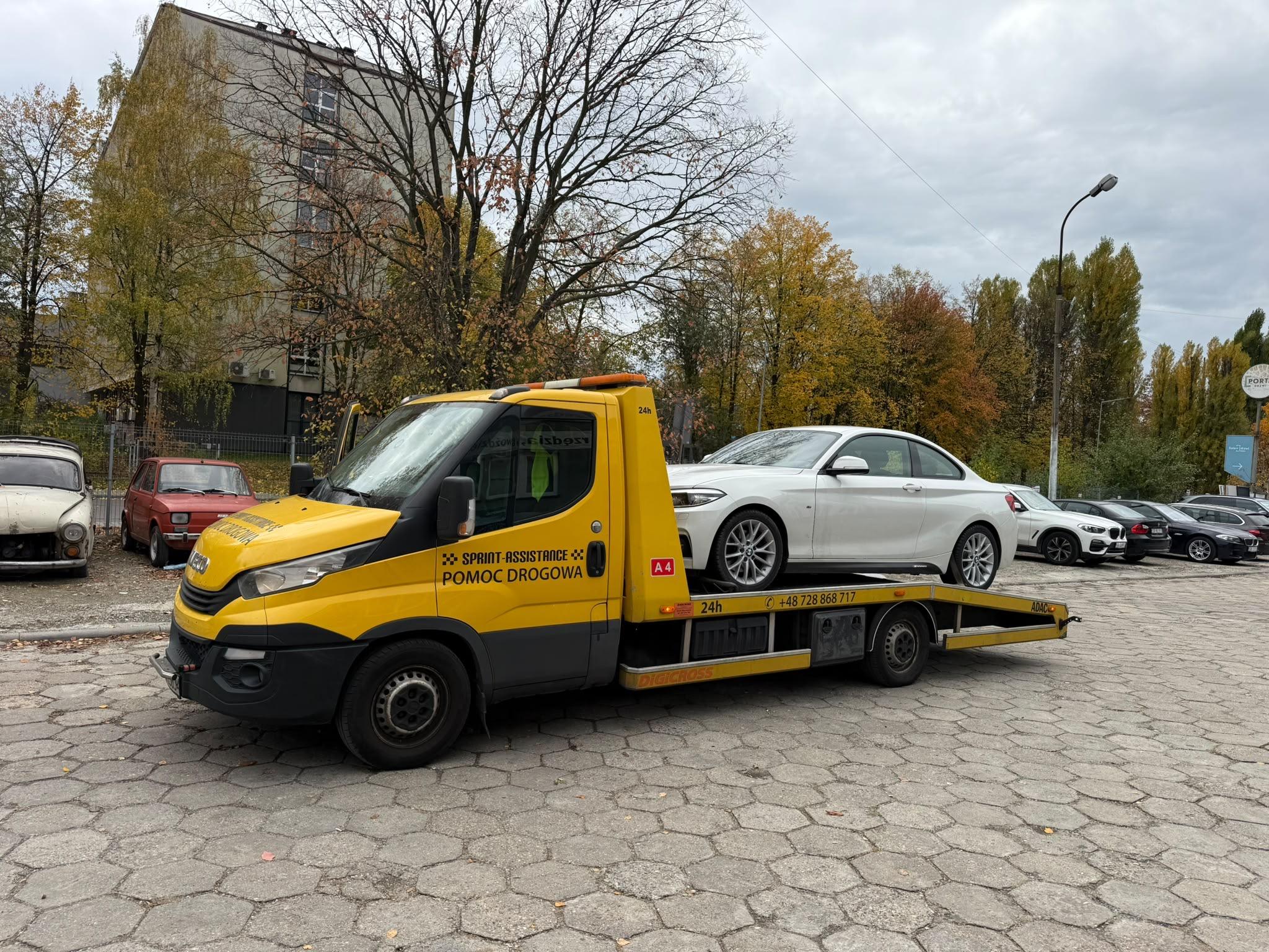 Laweta z białym BMW coupé na platformie, jesienny parking