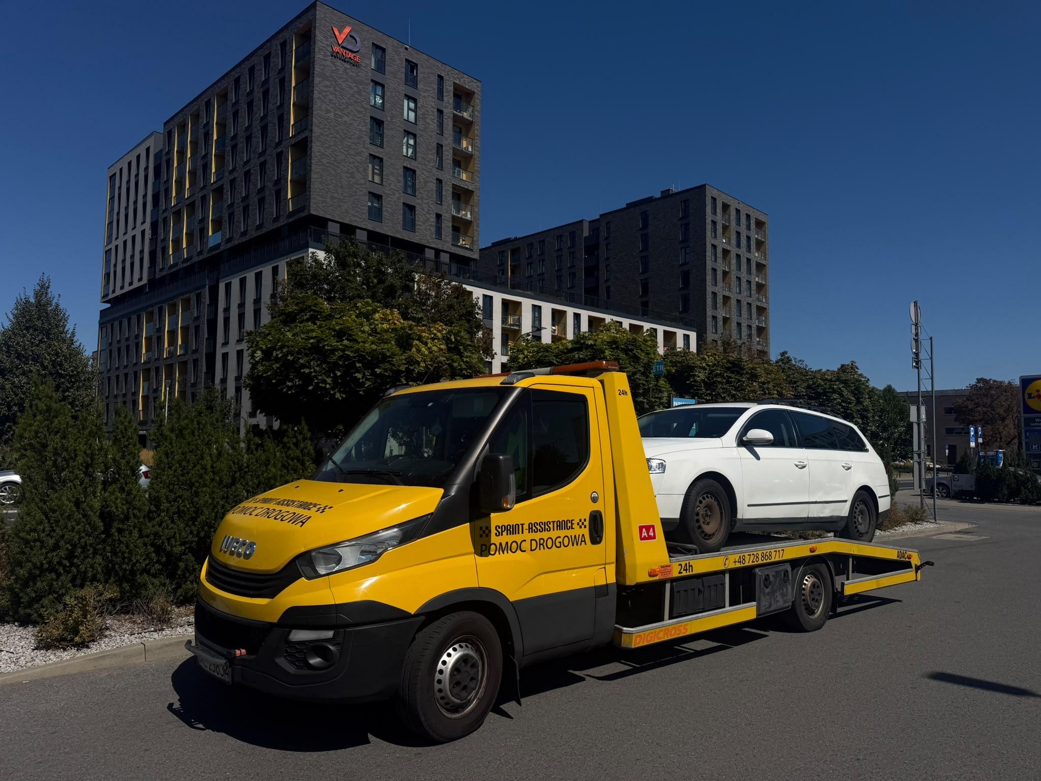 Laweta z białym kombi na platformie przed nowym osiedlem