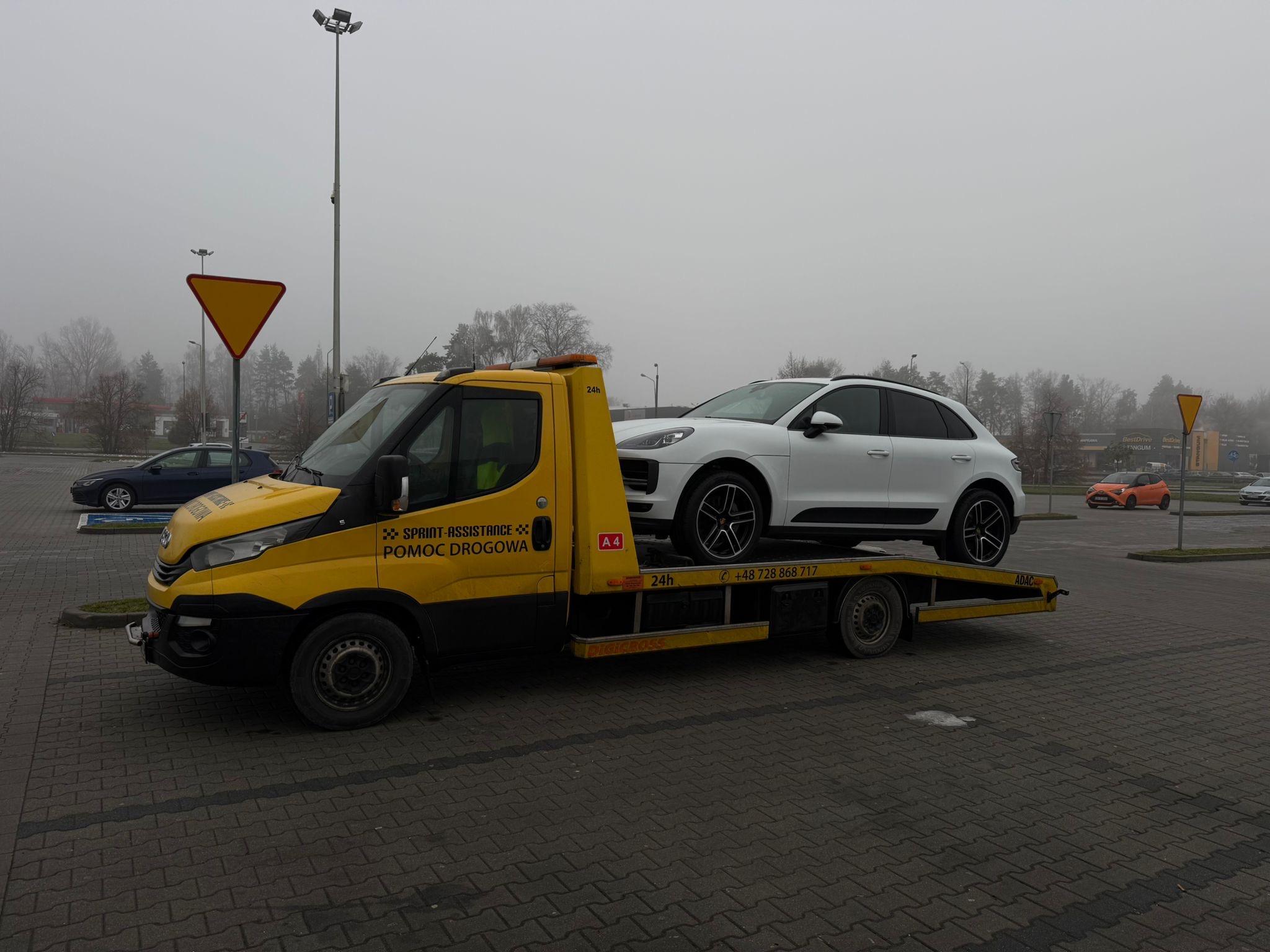 Laweta z białym SUV-em Porsche na platformie, parking we mgle