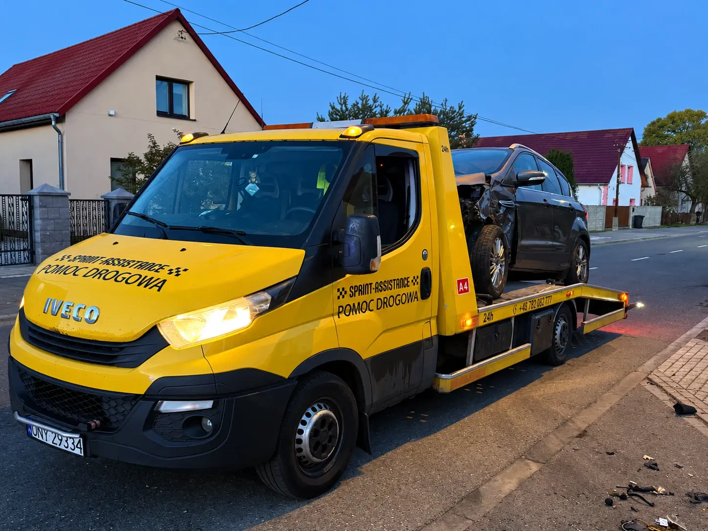 Laweta Iveco z rozbitym ciemnym SUV-em na platformie na ulicy między domami o zmierzchu