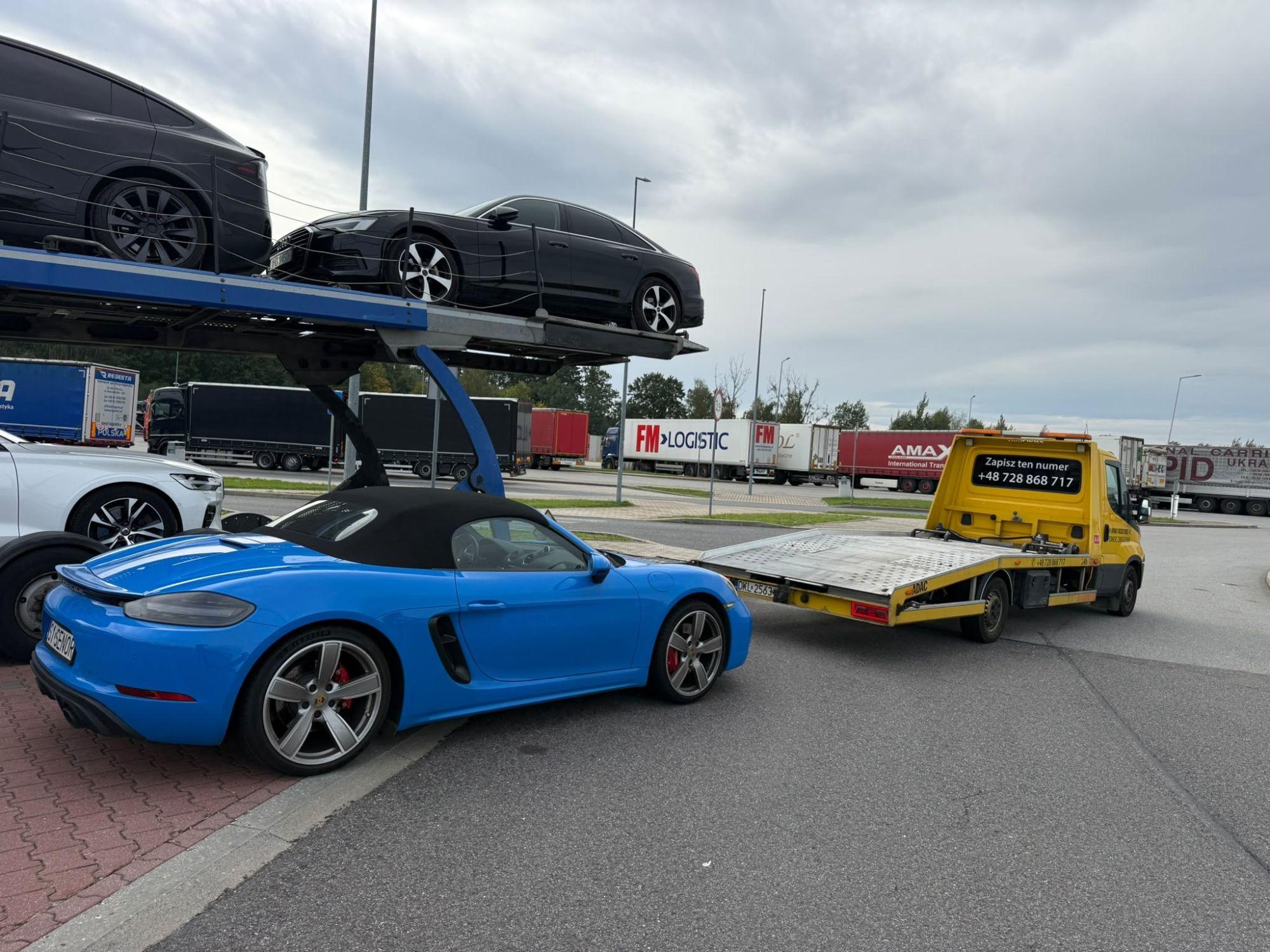 Niebieskie Porsche kabriolet przed lawetą z opuszczoną platformą na parkingu przy autostradzie, w tle autotransporter i ciężarówki